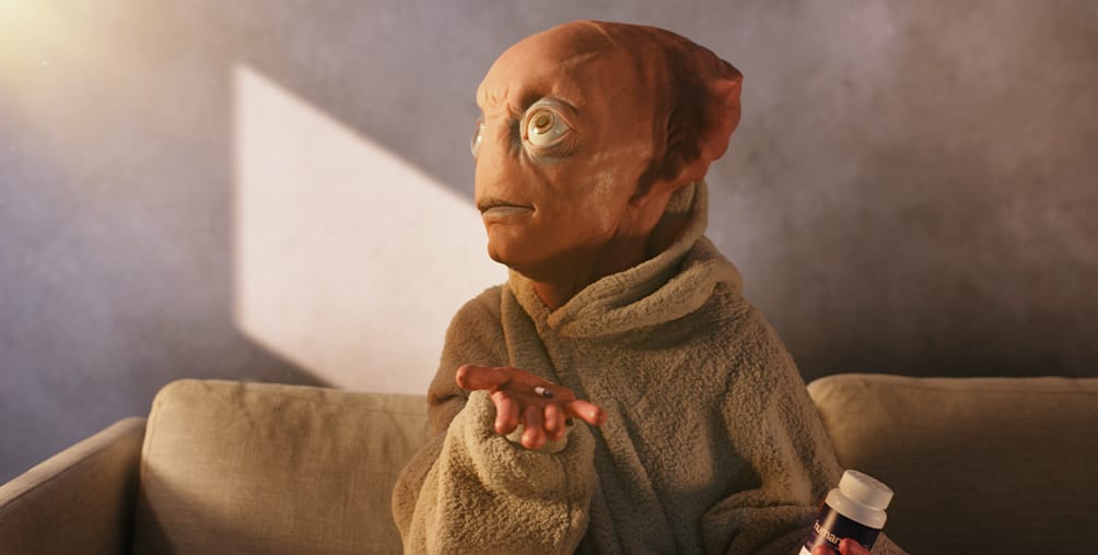 A person in a brown, alien-like mask and beige robe sits on a couch, holding a pill bottle in one hand and extending the other hand with pills, in a softly lit room.