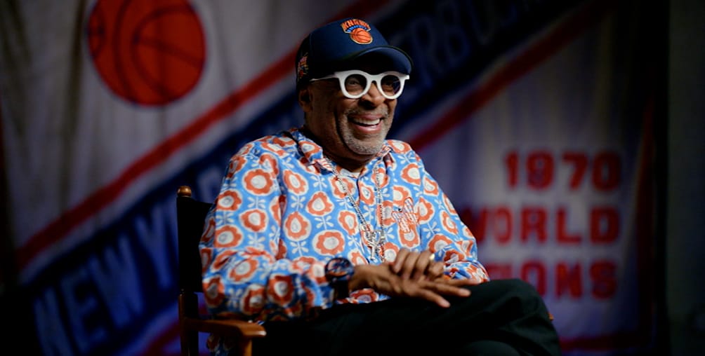 A man wearing a colorful patterned shirt, white glasses, and a New York Knicks cap smiles while sitting in front of a large New York Knicks 1970 World Champions banner.