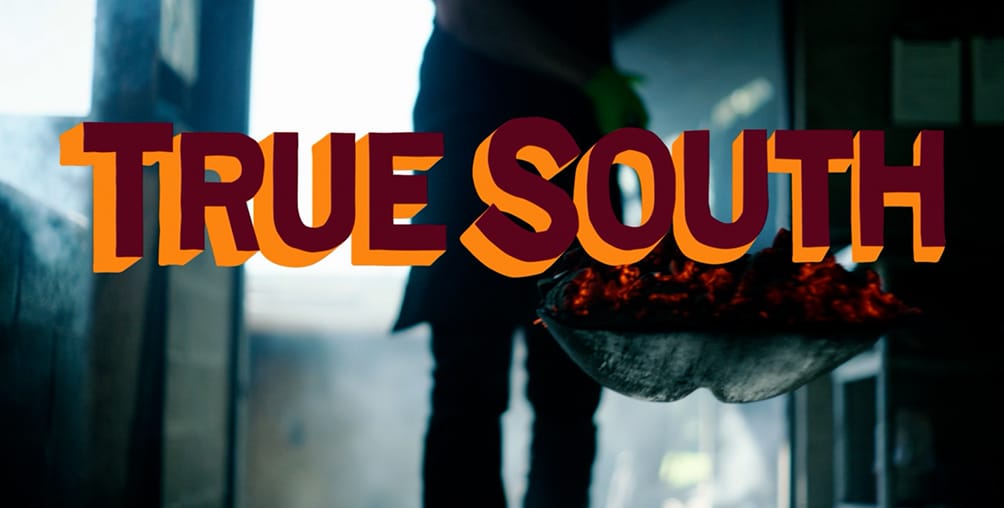 The image shows the words “TRUE SOUTH” in bold, colorful letters. Behind the text, a person stands holding a pan with glowing coals, with smoke and soft light in the background.